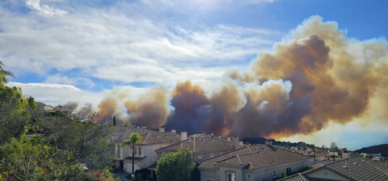 Large plume of wildfire smoke rising behind Southern California hillside homes with tile roofs, showing the threat of fire season to residential neighborhoods.
